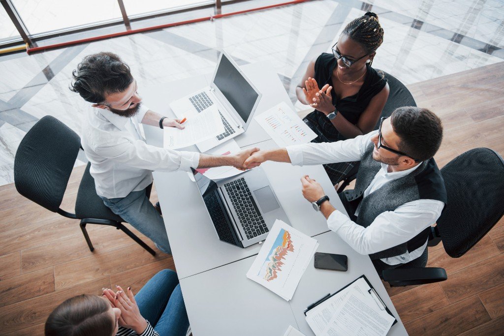 Business team celebrating a partnership handshake around a strategy table with laptops and analytics charts